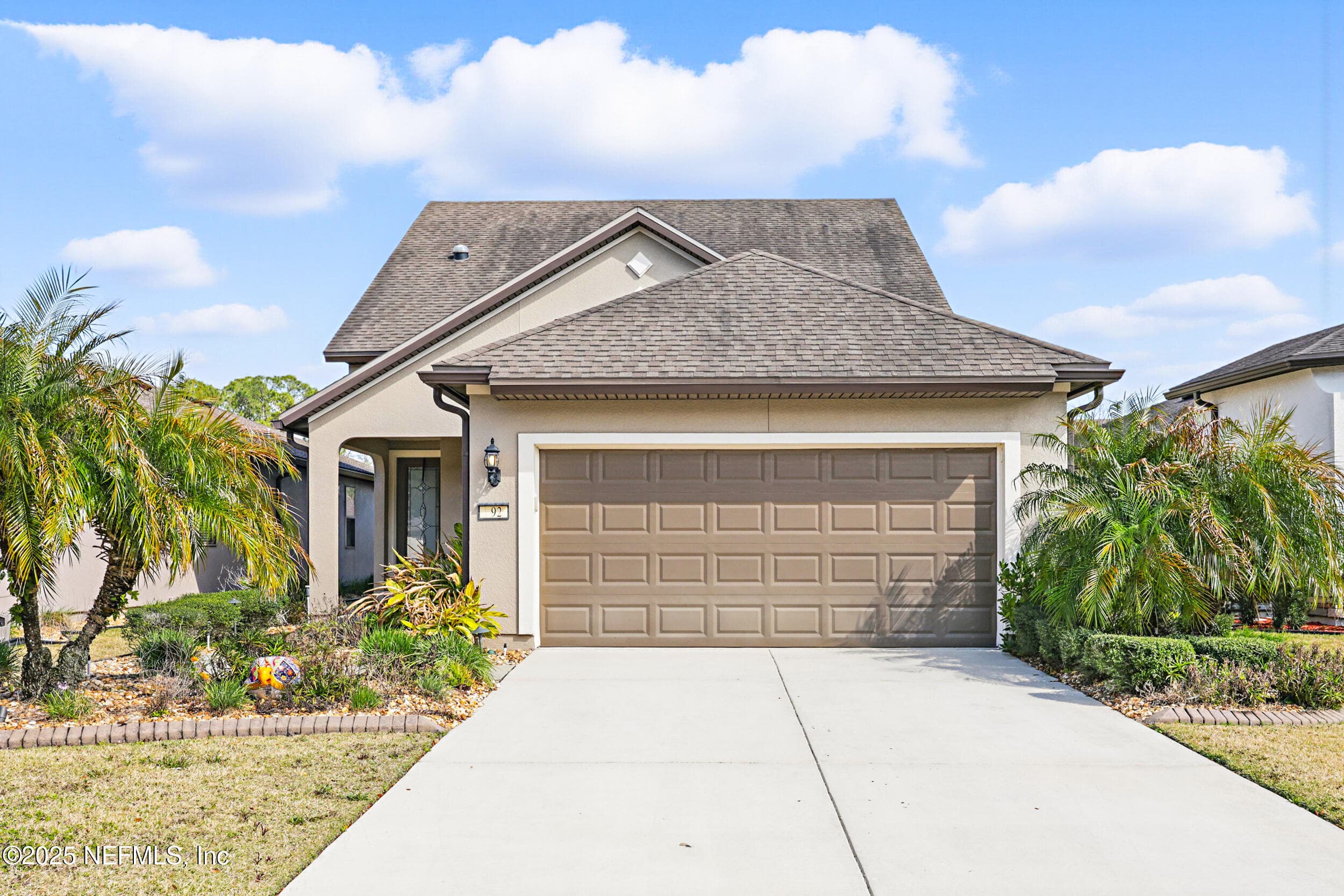 Front exterior (daytime) — Two-story stucco exterior with neutral paint, two-car garage with concrete driveway, covered entry with coach light fixture, and mature palm and tropical plantings on both sides. — 92 CANOPY OAK Lane, Ponte Vedra, FL 32081