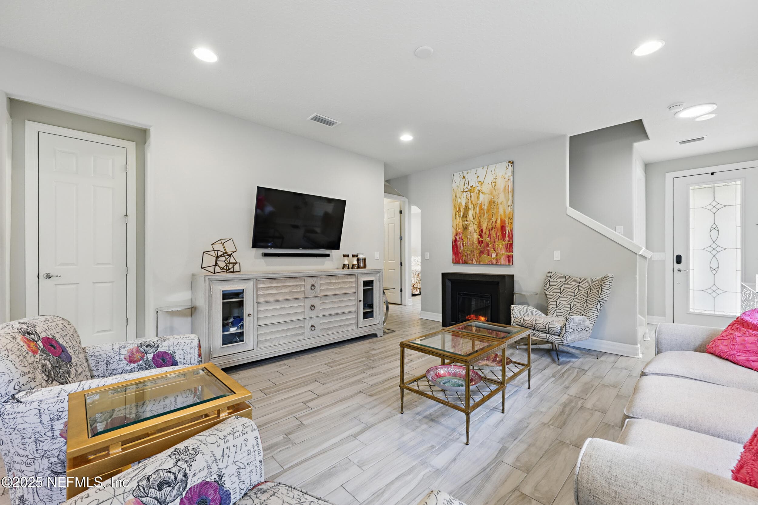 Living room (alternate angle) — Living room with electric fireplace, wall-mounted TV, wood-look tile flooring, recessed lighting, and open sightline to the foyer staircase and decorative leaded glass front door. Coat closet adjacent to living area. — 92 CANOPY OAK Lane, Ponte Vedra, FL 32081