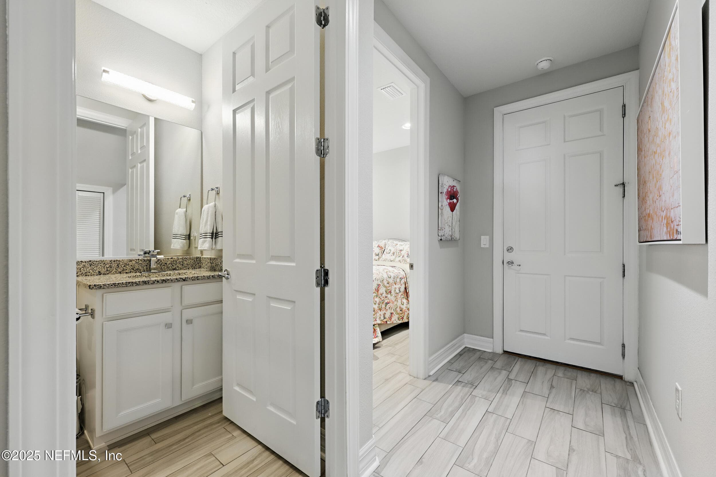 First-floor hallway — Hallway connecting the second bedroom, guest bathroom, and garage entry with wood-look tile flooring. Guest bath features granite countertop, white shaker cabinetry, and framed mirror. — 92 CANOPY OAK Lane, Ponte Vedra, FL 32081