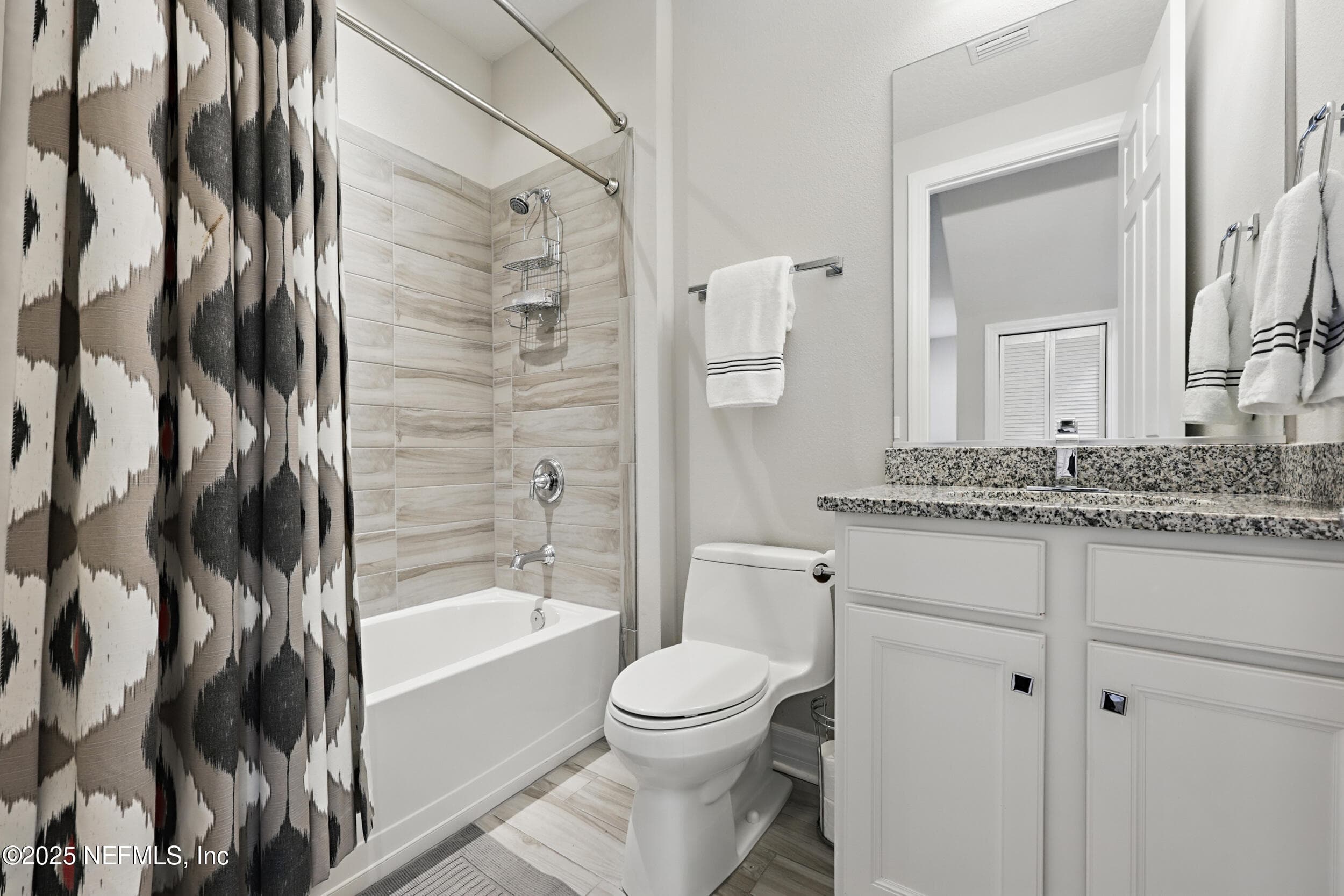 Guest bathroom — Full guest bath with tub-shower combo, wood-look tile surround, granite vanity countertop, white shaker cabinet, framed mirror, and chrome fixtures. Curved shower rod and wood-look tile flooring. — 92 CANOPY OAK Lane, Ponte Vedra, FL 32081