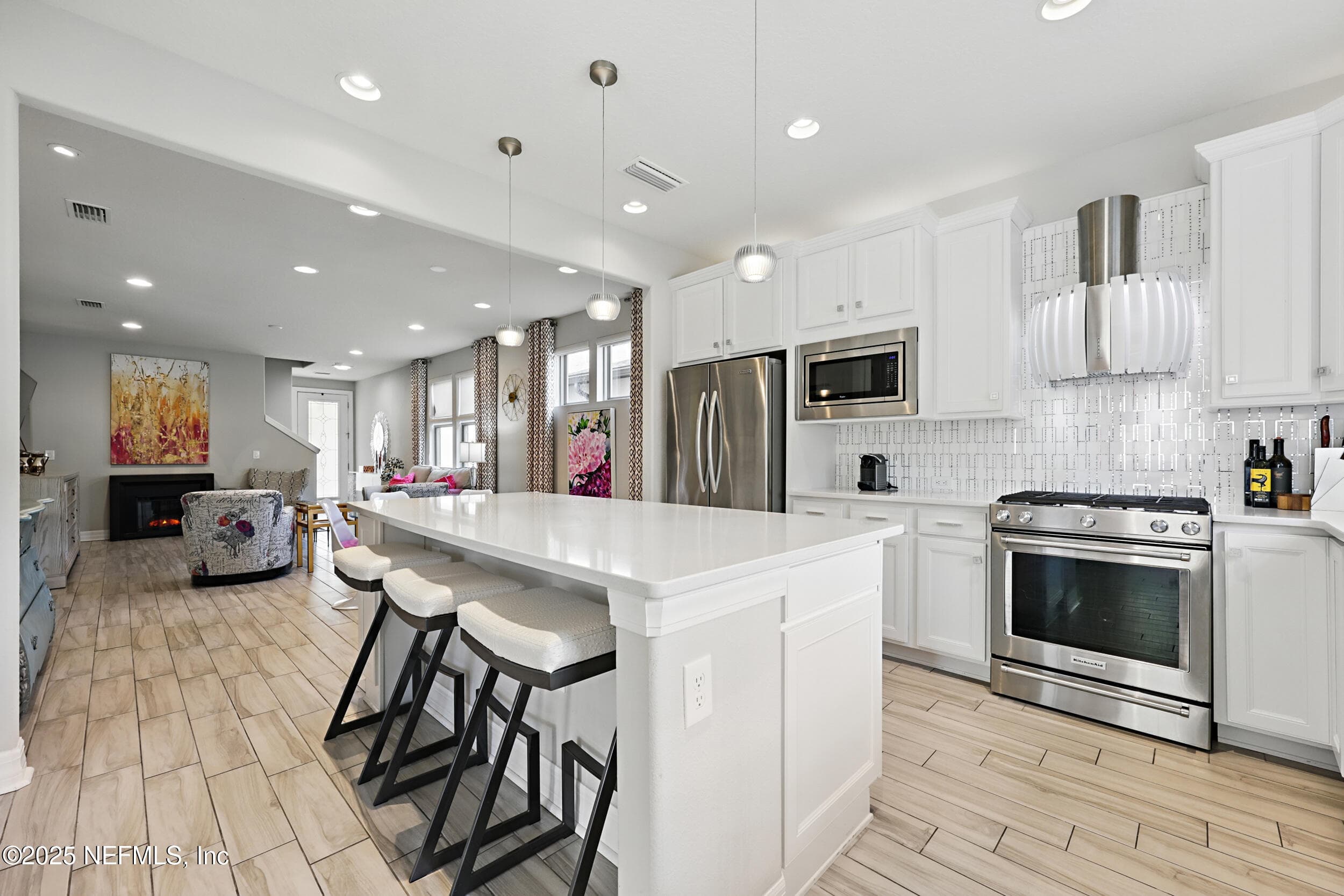 Kitchen (toward living and dining) — Open-concept kitchen with island seating for three, KitchenAid gas range, custom stainless steel and glass vent hood, white quartz countertops, geometric tile backsplash, and sightline through to the dining area, living room, and fireplace. — 92 CANOPY OAK Lane, Ponte Vedra, FL 32081