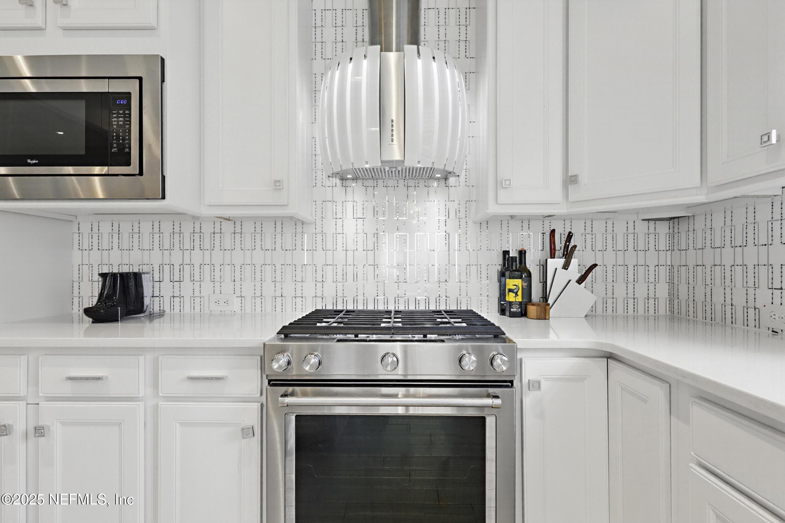 Kitchen (range and vent hood detail) — KitchenAid stainless steel gas range with custom wall-mounted vent hood, geometric mosaic tile backsplash, white quartz countertops, white shaker cabinetry, and built-in Whirlpool microwave. — 92 CANOPY OAK Lane, Ponte Vedra, FL 32081
