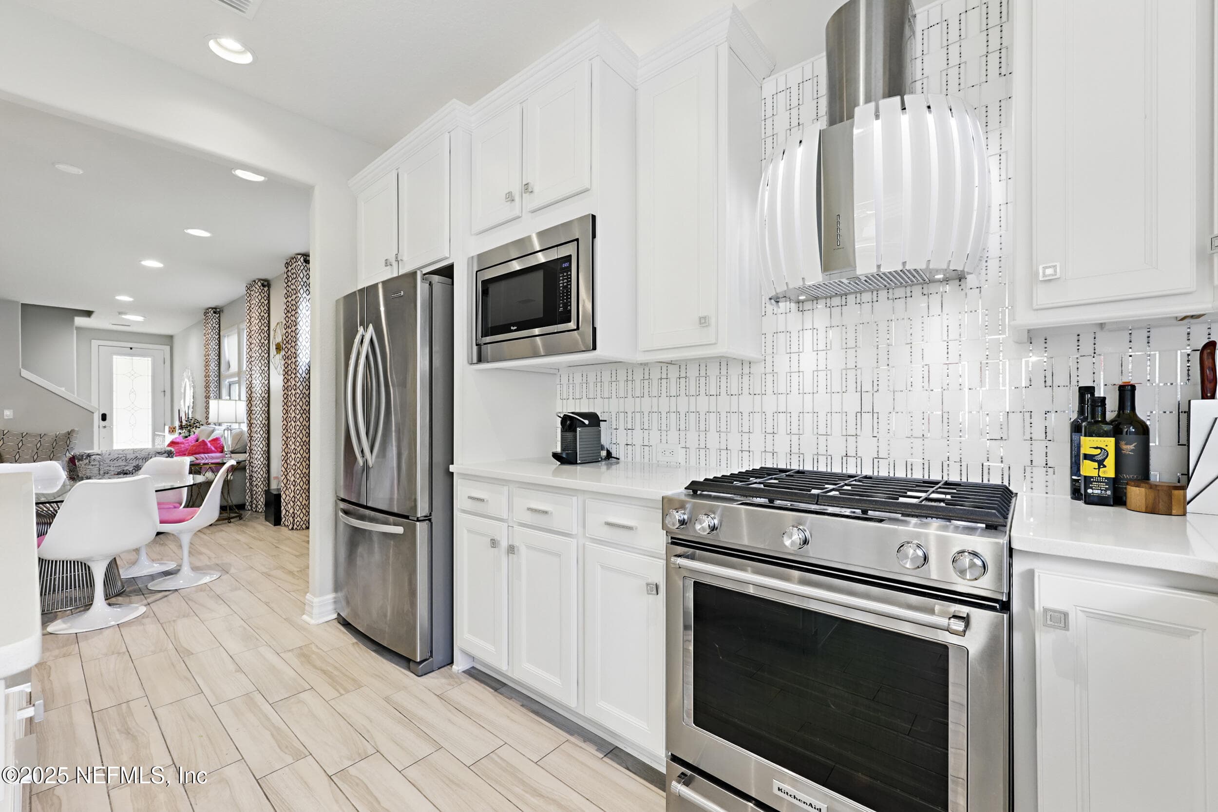 Kitchen (appliance wall) — KitchenAid gas range, stainless steel French door refrigerator, built-in microwave, custom vent hood, geometric tile backsplash, white shaker cabinetry with crown molding, and wood-look tile flooring. Open sightline through to the dining and living areas. — 92 CANOPY OAK Lane, Ponte Vedra, FL 32081