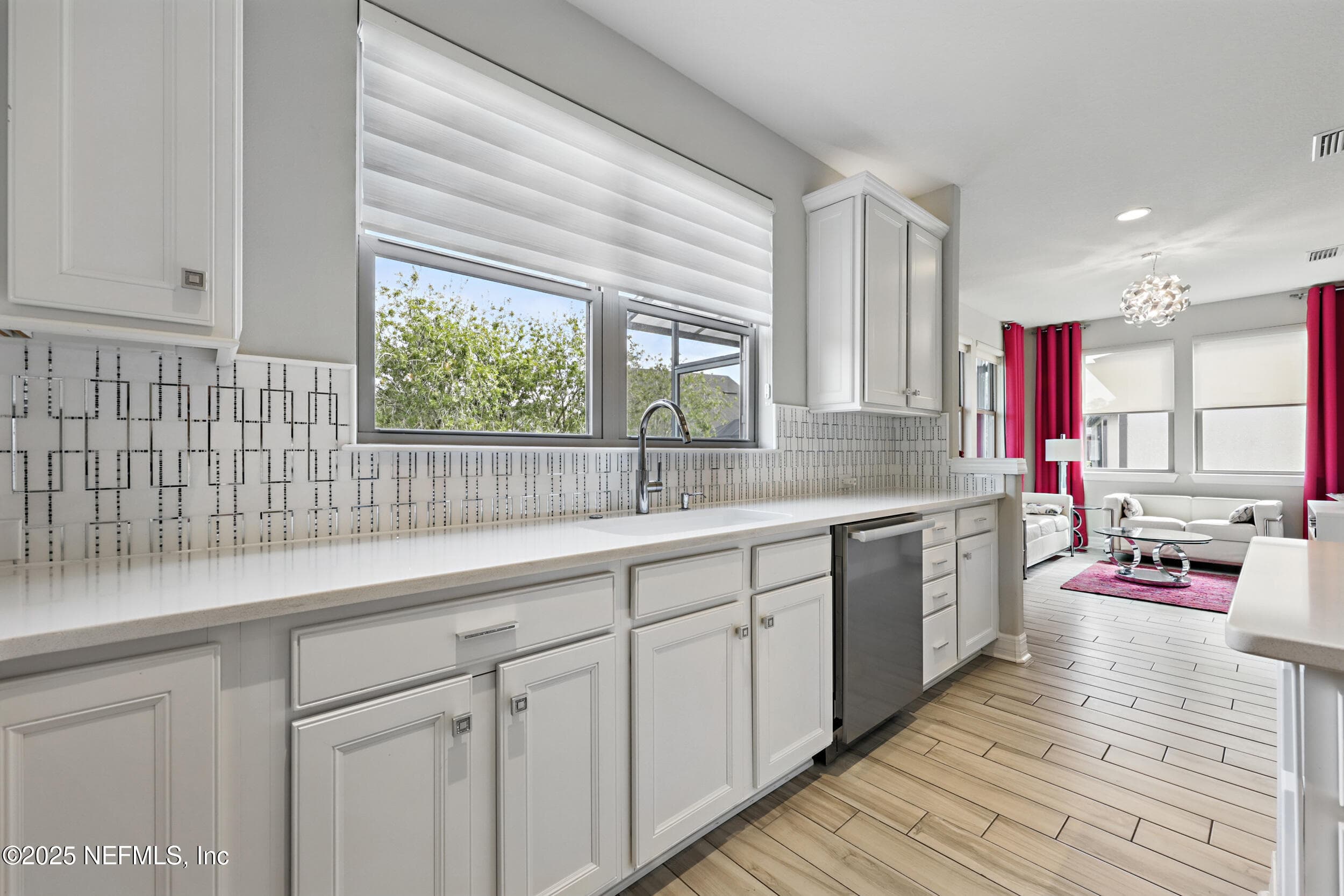 Kitchen (sink wall) — White quartz countertop with undermount sink, high-arc chrome pull-down faucet, stainless steel dishwasher, geometric tile backsplash, window with custom blinds above the sink, and white shaker cabinetry. View through to the sitting room with decorative chandelier. — 92 CANOPY OAK Lane, Ponte Vedra, FL 32081