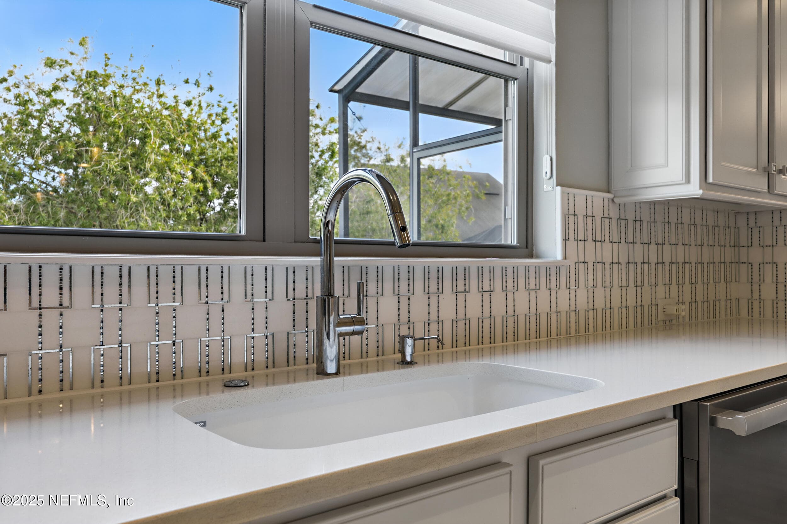 Kitchen (sink close-up) — Undermount white sink with chrome high-arc pull-down faucet, white quartz countertop, geometric mosaic tile backsplash, and window with tree-lined view. Stainless steel dishwasher visible to the right. — 92 CANOPY OAK Lane, Ponte Vedra, FL 32081