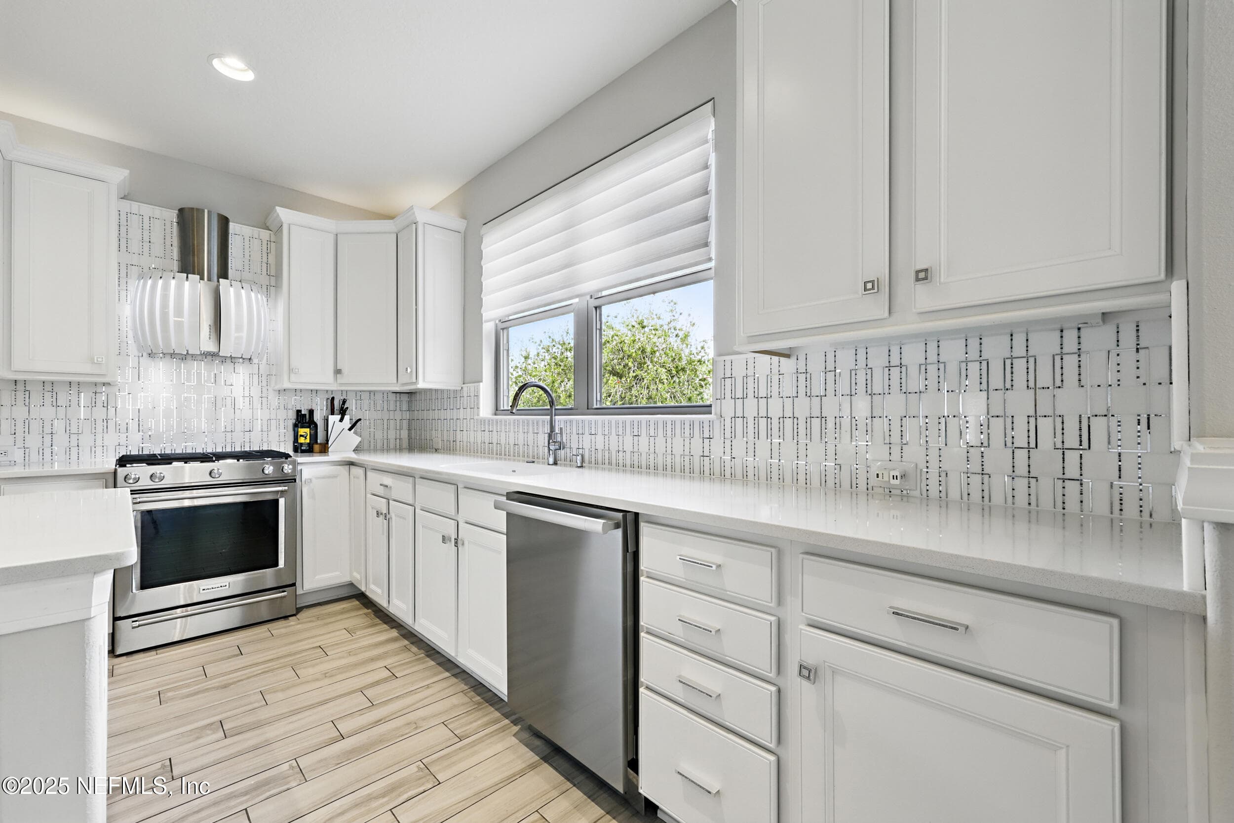 Kitchen (perimeter counters) — L-shaped kitchen layout with KitchenAid gas range, stainless steel dishwasher, custom vent hood, white quartz countertops, geometric tile backsplash, window with custom blinds above the sink, and white shaker cabinetry throughout. — 92 CANOPY OAK Lane, Ponte Vedra, FL 32081