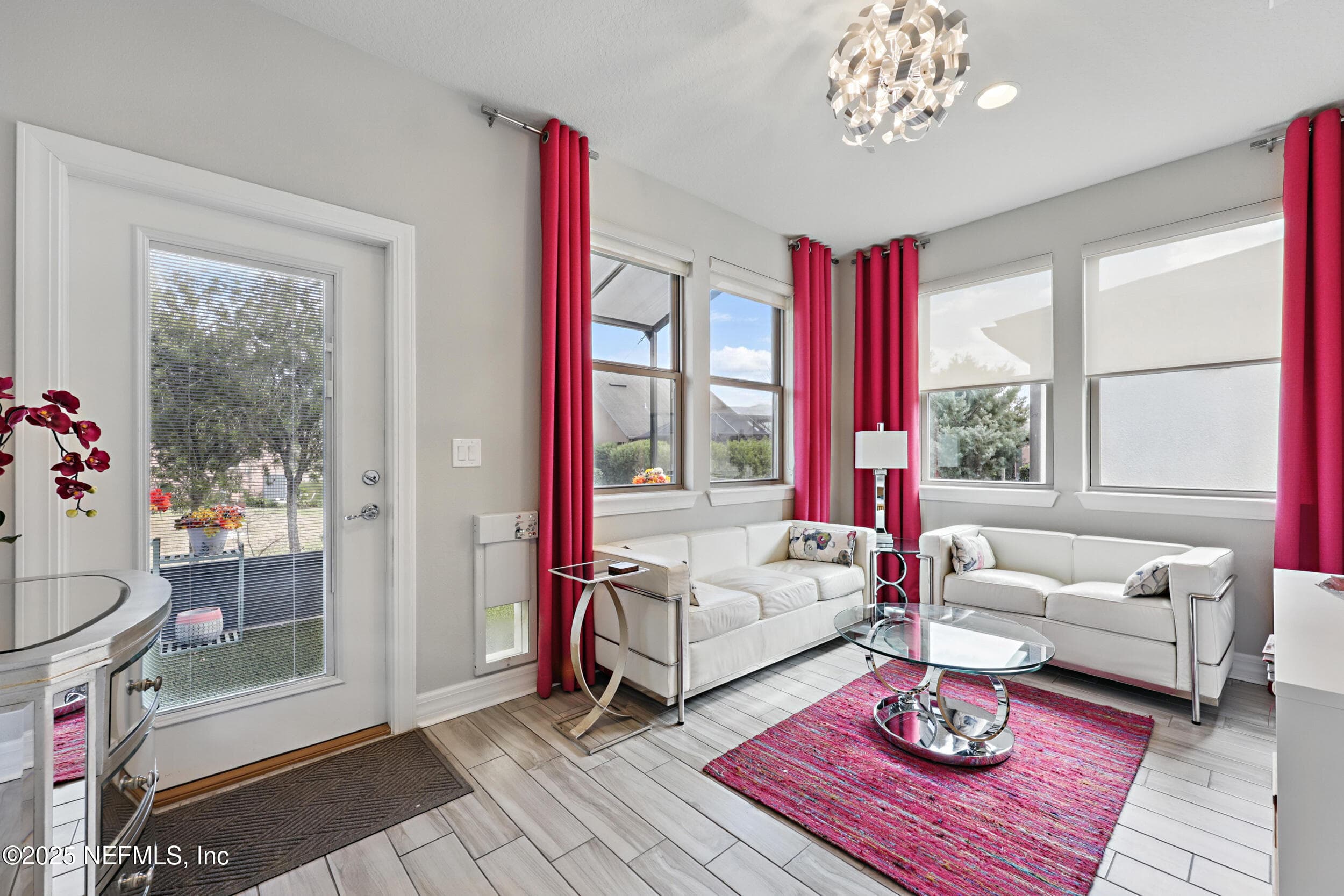 Sitting room/morning room — First-floor sitting room off the kitchen with glass patio door leading to the screened lanai, multiple windows with custom curtain panels, decorative modern chandelier, wood-look tile flooring, and natural light throughout. — 92 CANOPY OAK Lane, Ponte Vedra, FL 32081