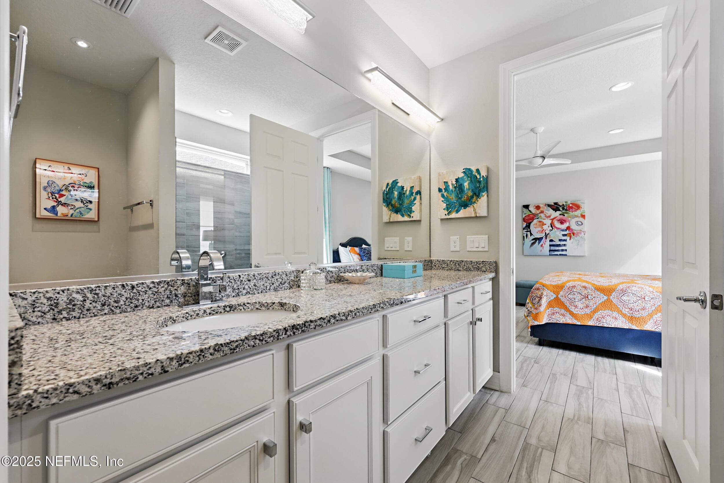 Primary bathroom (double vanity detail) — Primary en-suite with granite double-sink vanity, white shaker cabinetry, brushed nickel hardware, full-width frameless mirror, and open doorway to the primary bedroom with tray ceiling. — 92 CANOPY OAK Lane, Ponte Vedra, FL 32081