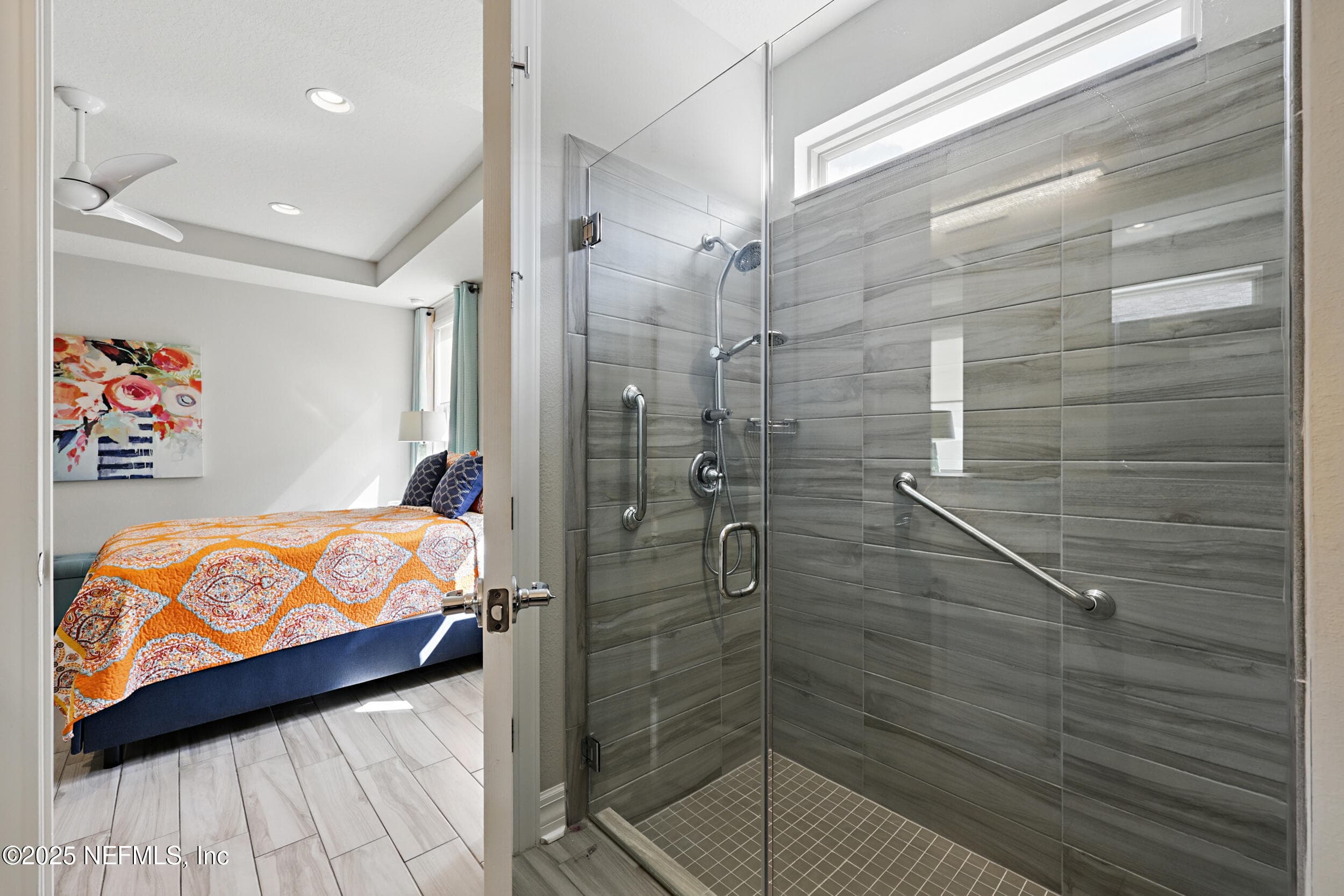 Primary bathroom (walk-in shower detail) — Frameless glass walk-in shower with floor-to-ceiling gray wood-look tile, built-in shower niche, handheld showerhead on slide bar, grab bars, chrome fixtures, and transom window for natural light. — 92 CANOPY OAK Lane, Ponte Vedra, FL 32081