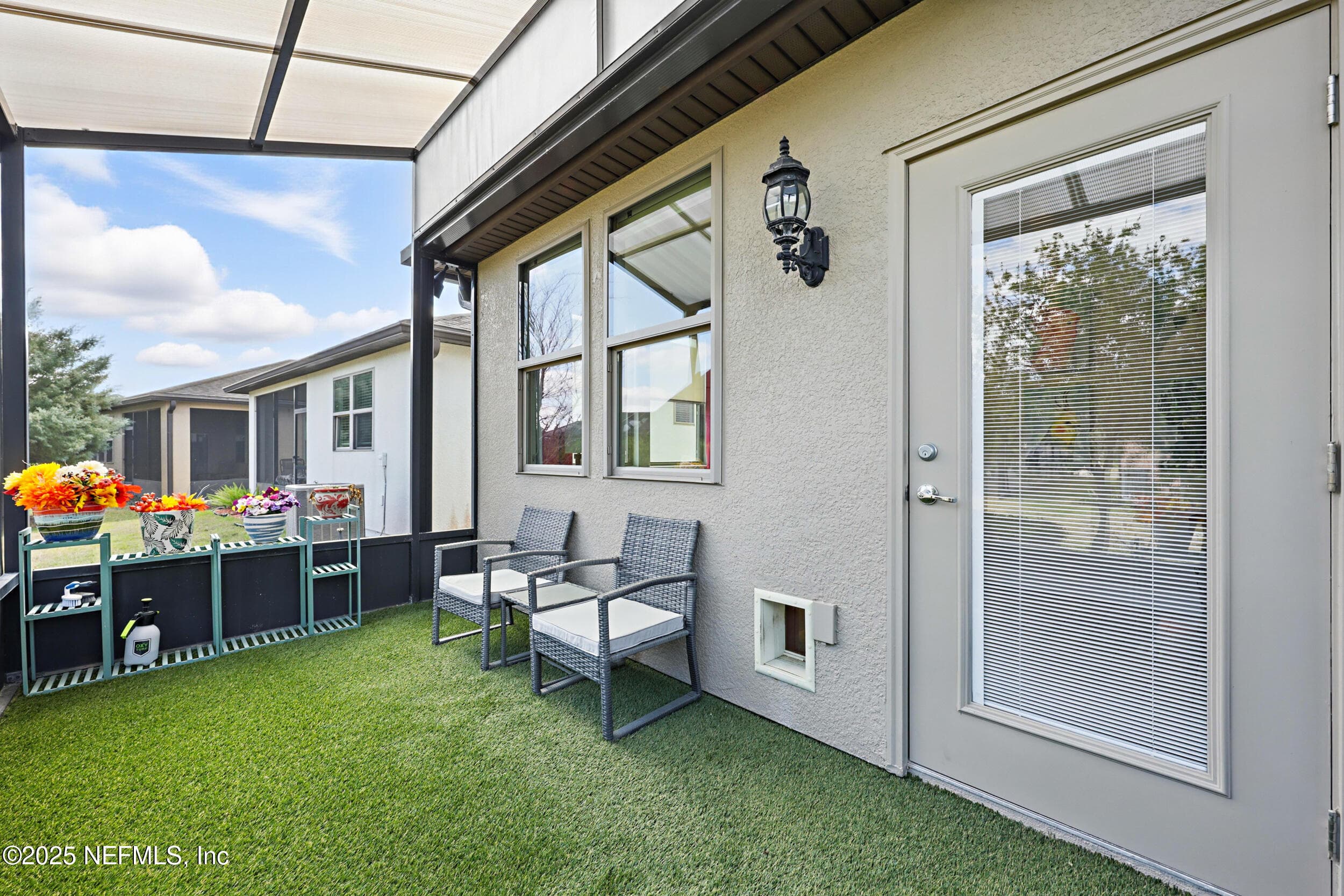Screened lanai (seating area) — Screened lanai with artificial turf, wicker seating area, coach light fixture, glass patio door entry from the sitting room, pet door access, and covered roof structure with tree-lined backyard view. — 92 CANOPY OAK Lane, Ponte Vedra, FL 32081