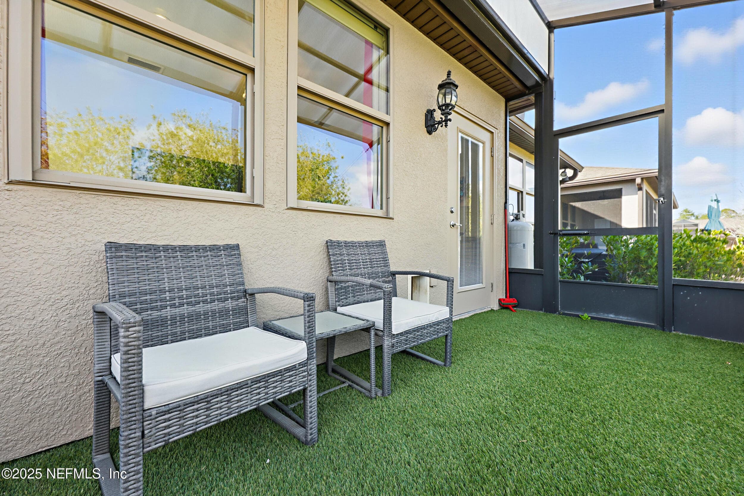 Screened lanai (seating detail) — Screened lanai with artificial turf, wicker bistro seating set, coach light fixture, glass patio door, and windows overlooking the sitting room. Covered roof provides shade with views of the landscaped backyard. — 92 CANOPY OAK Lane, Ponte Vedra, FL 32081