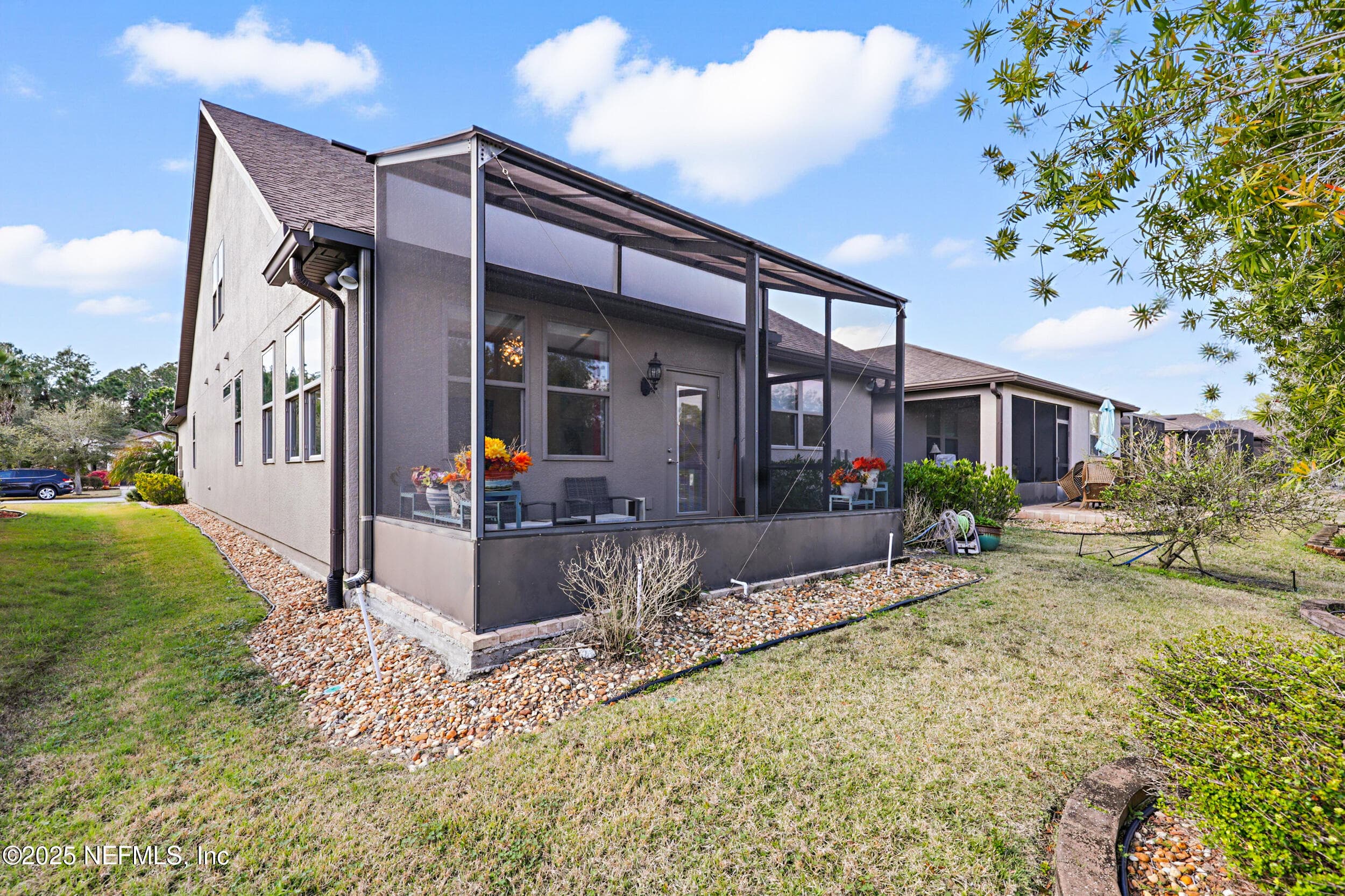 Rear exterior (wide angle) — Two-story stucco exterior with full screened lanai, river rock landscape bed, backyard with mature plantings, and side yard with paver border. Architectural shingle roof and coach light fixture at the lanai entry. — 92 CANOPY OAK Lane, Ponte Vedra, FL 32081