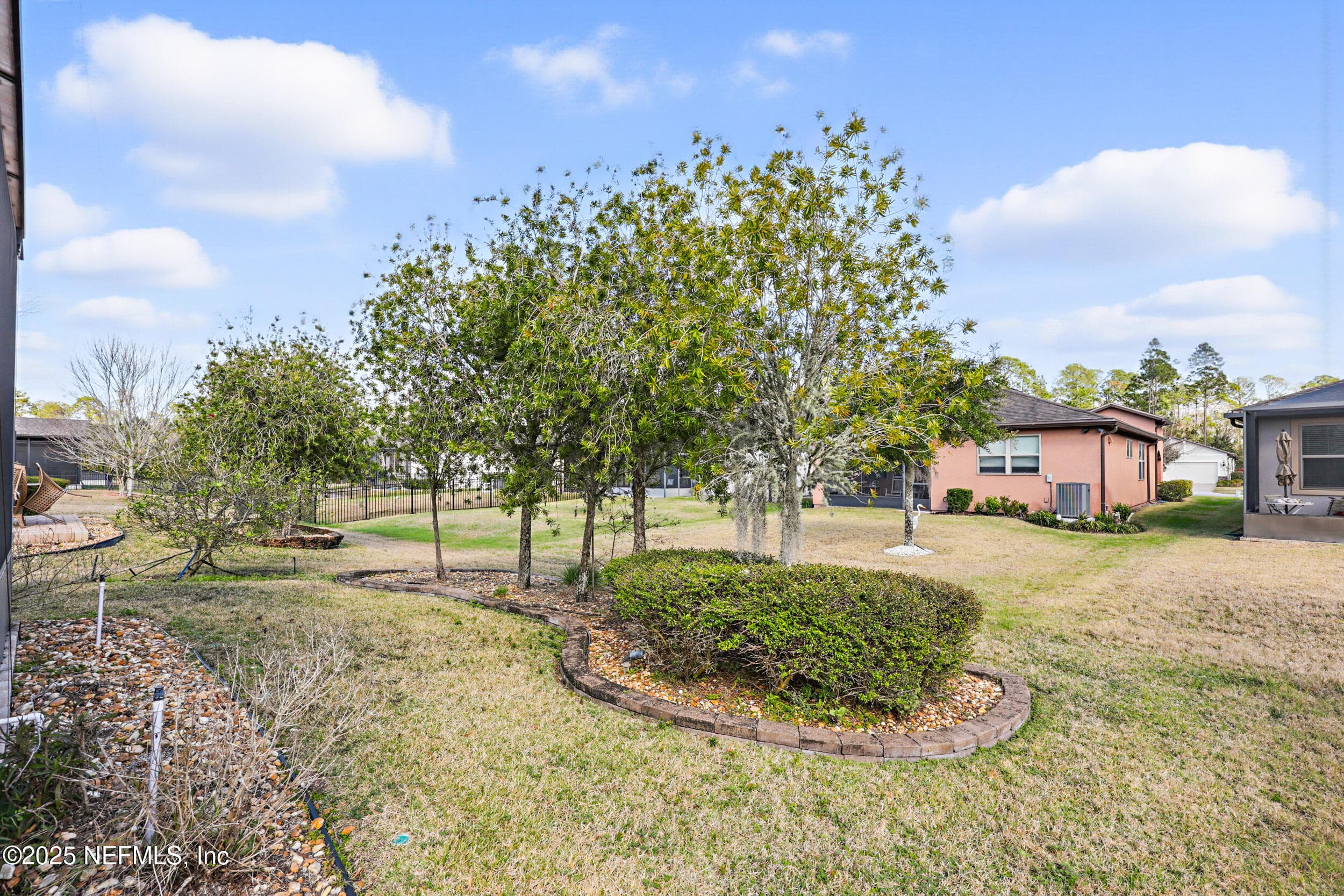 Backyard — Landscaped backyard with mature trees, paver-edged planting beds, manicured lawn, and views of the Del Webb Ponte Vedra community. Iron perimeter fencing visible along the rear property line. — 92 CANOPY OAK Lane, Ponte Vedra, FL 32081