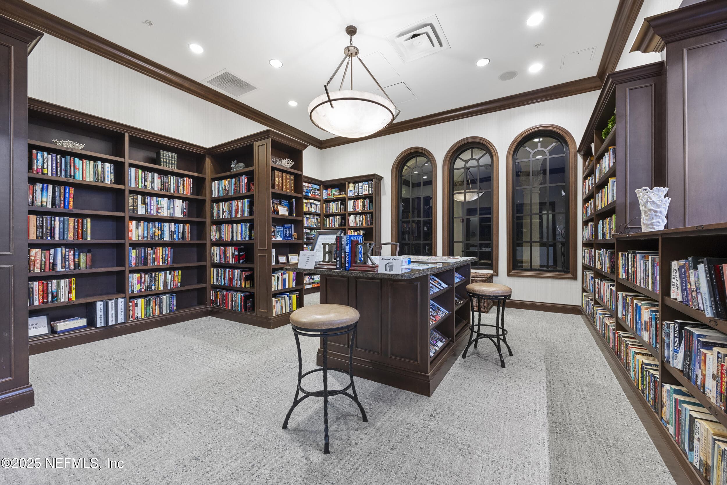 Anastasia Clubhouse library — Resident library inside the Anastasia Clubhouse with floor-to-ceiling dark wood built-in bookshelves, center island with granite top and barstool seating, arched windows, pendant chandelier, and carpeted flooring. — 92 CANOPY OAK Lane, Ponte Vedra, FL 32081