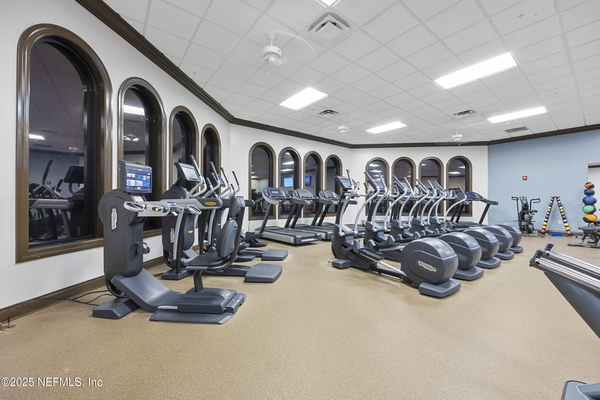 Anastasia Clubhouse fitness center (cardio) — Cardio room with Technogym ellipticals, treadmills, and recumbent bikes with touchscreen displays, arched mirrored windows, and open layout inside the Anastasia Clubhouse at Del Webb Ponte Vedra. — 92 CANOPY OAK Lane, Ponte Vedra, FL 32081