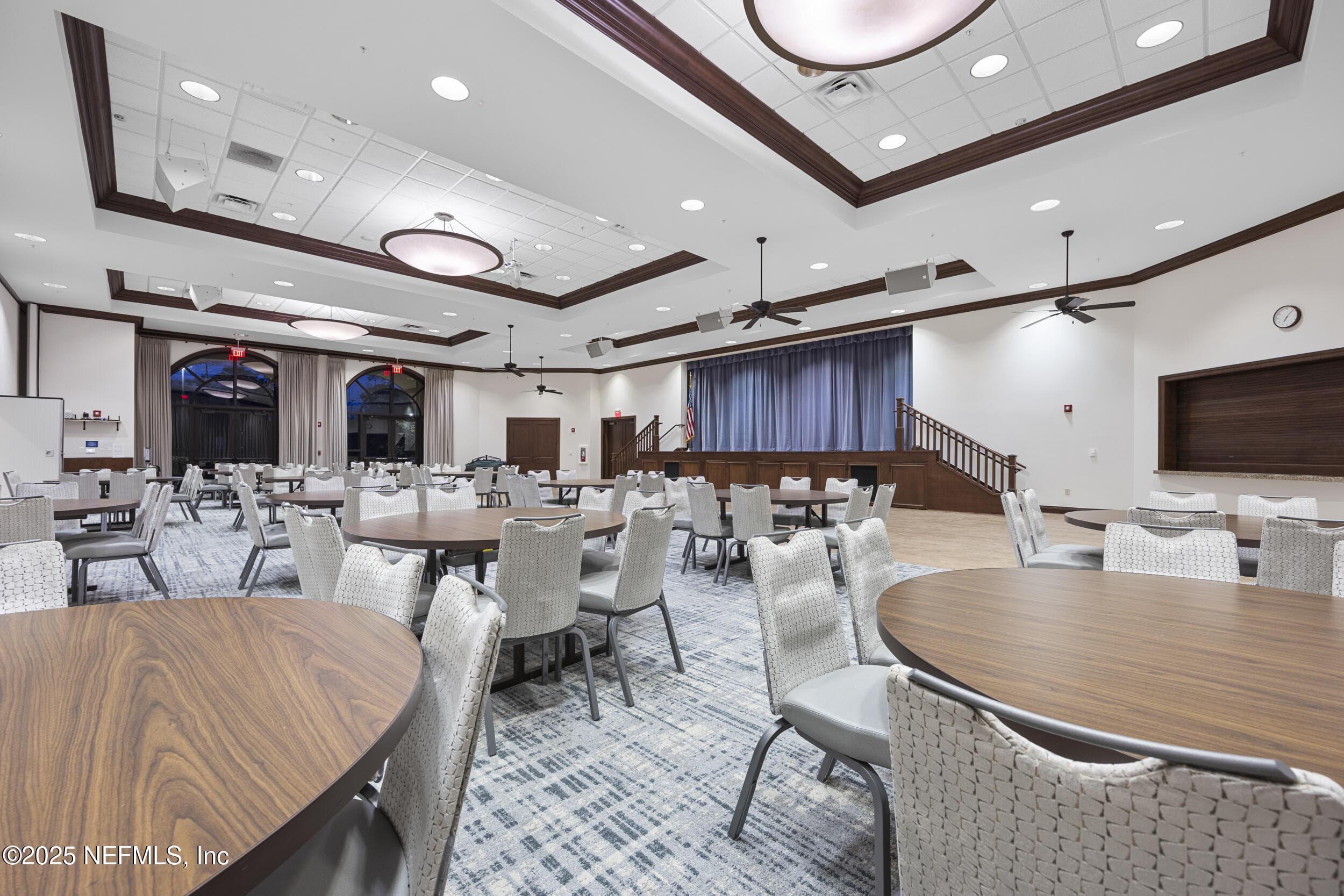 Anastasia Clubhouse ballroom — Ballroom inside the Anastasia Clubhouse with banquet seating, tray ceiling with crown molding, arched windows, ceiling fans, wall-mounted TV, and divider curtain for flexible event configurations at Del Webb Ponte Vedra. — 92 CANOPY OAK Lane, Ponte Vedra, FL 32081
