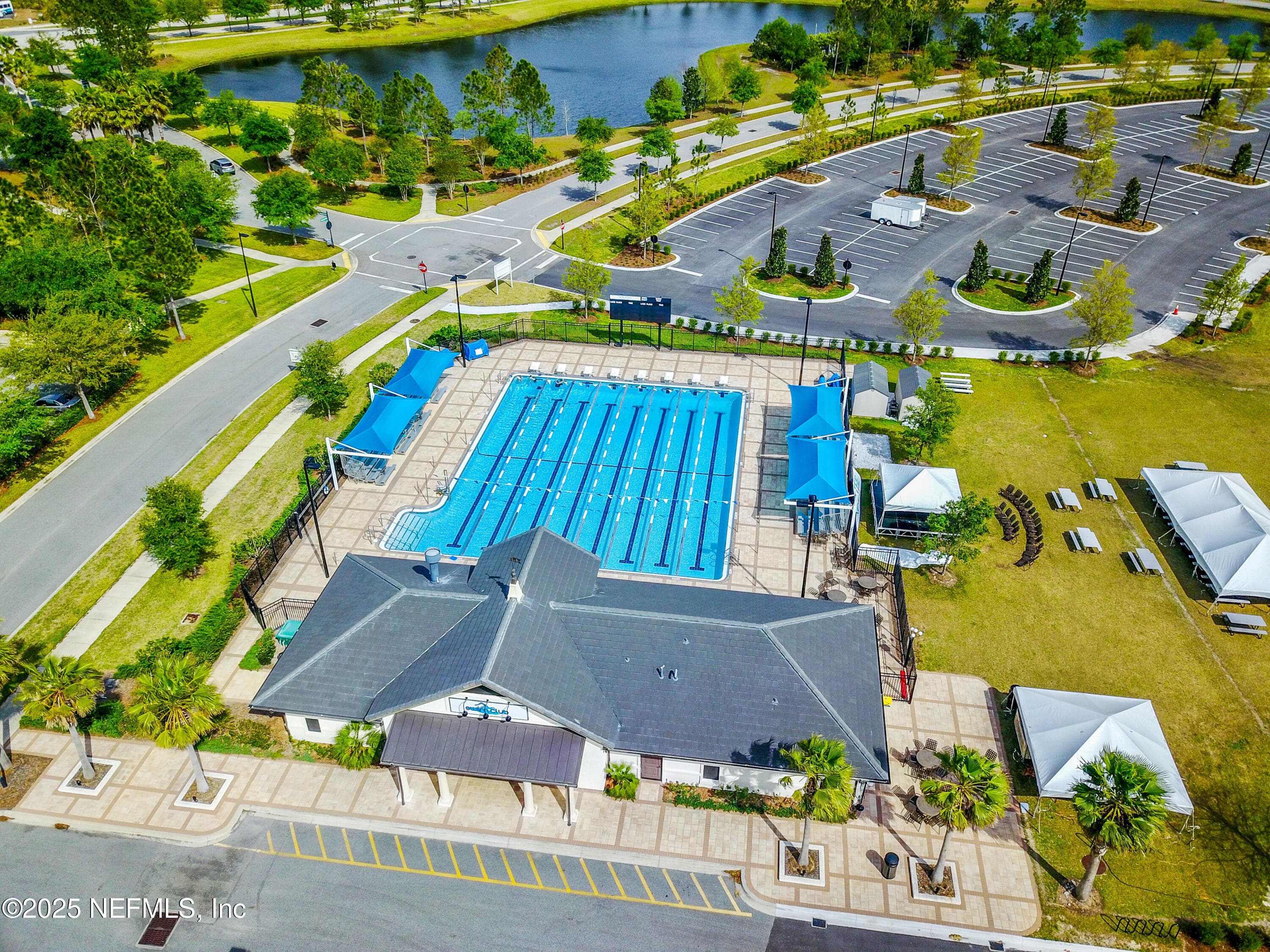 Nocatee lap pool and swim club (aerial) — Aerial view of the Nocatee swim club with competition-style lap pool, lane dividers, shade structures, clubhouse building, lakeside setting, and event lawn with tent pavilions in Ponte Vedra, FL. — 92 CANOPY OAK Lane, Ponte Vedra, FL 32081